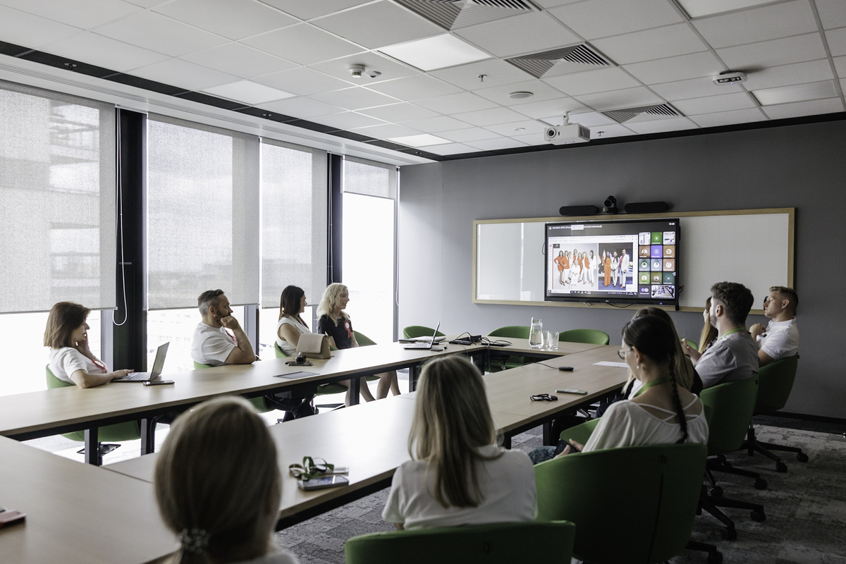 Ten Leroy Merlin team members sit in a conference room during an online meeting. On the screen is a presentation with female speakers. On the table a glass pitcher and glasses.